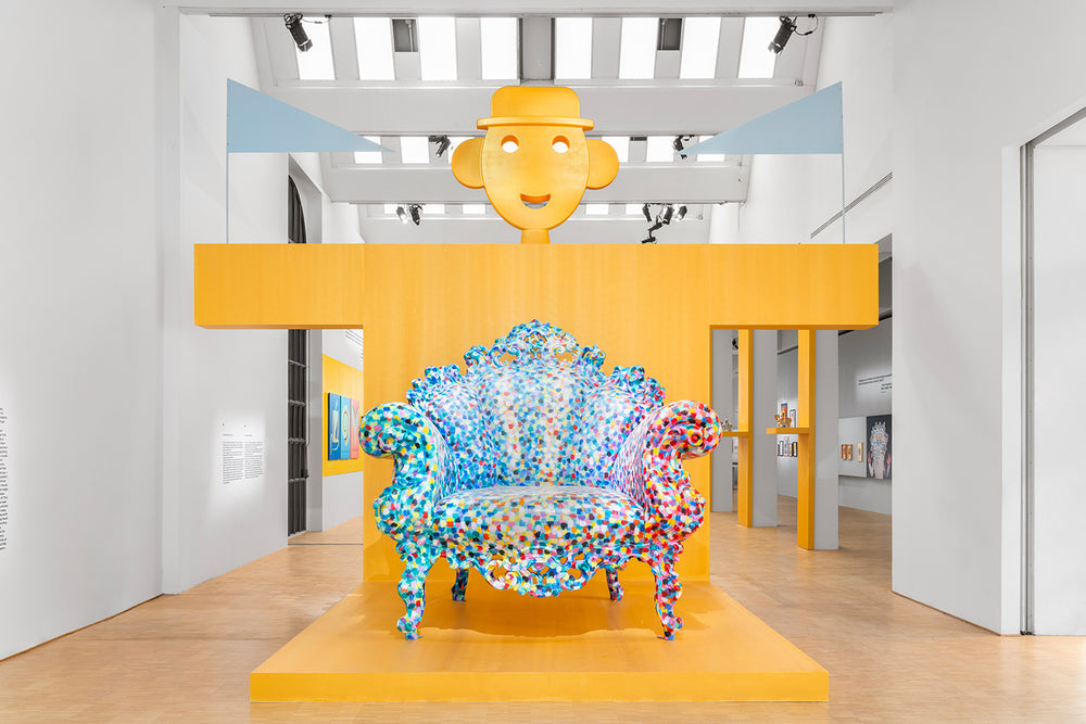 Vibrant, polka-dotted armchair under a cheerful yellow smiley face sculpture in a bright gallery space.