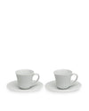 Two white ceramic cups with saucers by Alessi, featuring a simple, elegant curved design, presented at a slight angle.