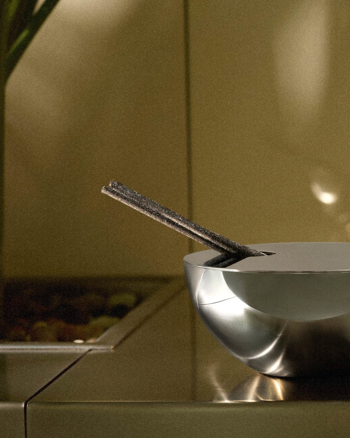 Silver Rocc fragrance diffuser by Alessi, a sleek metallic bowl holding dark incense sticks against a warm, grainy background.