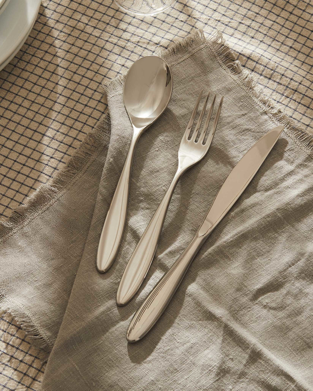 Polished stainless steel table fork by Alessi, alongside a matching spoon and knife on a fringed linen napkin and plaid tablecloth.