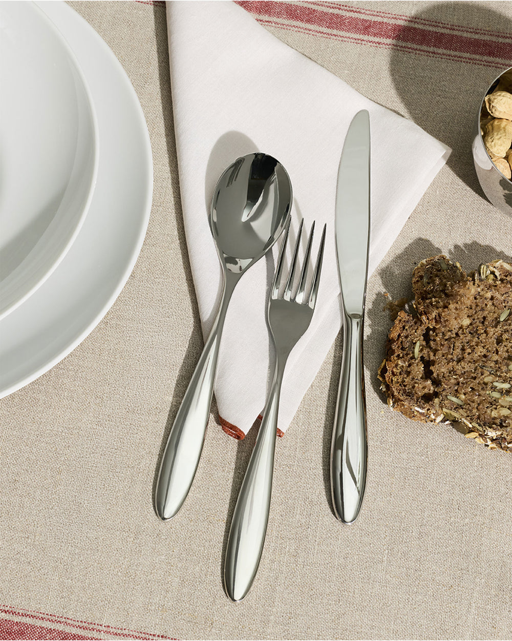 Polished stainless steel Mami table fork by Alessi, resting on a white napkin with other cutlery and a striped tablecloth.