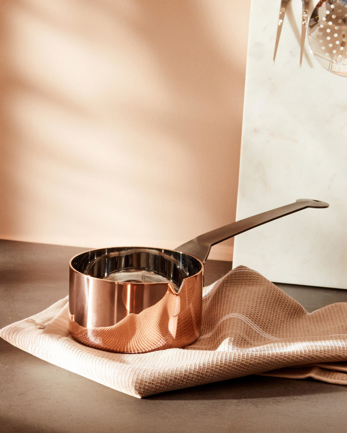 Polished copper saucepan by Alessi with a flat black handle and pouring spout, on a beige waffle towel.