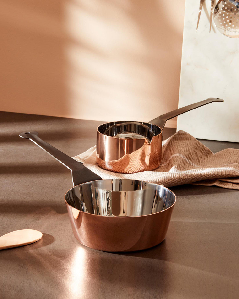 Two shiny copper saucepans by Alessi with matte black handles, one smaller with a pouring spout, on a dark kitchen counter.