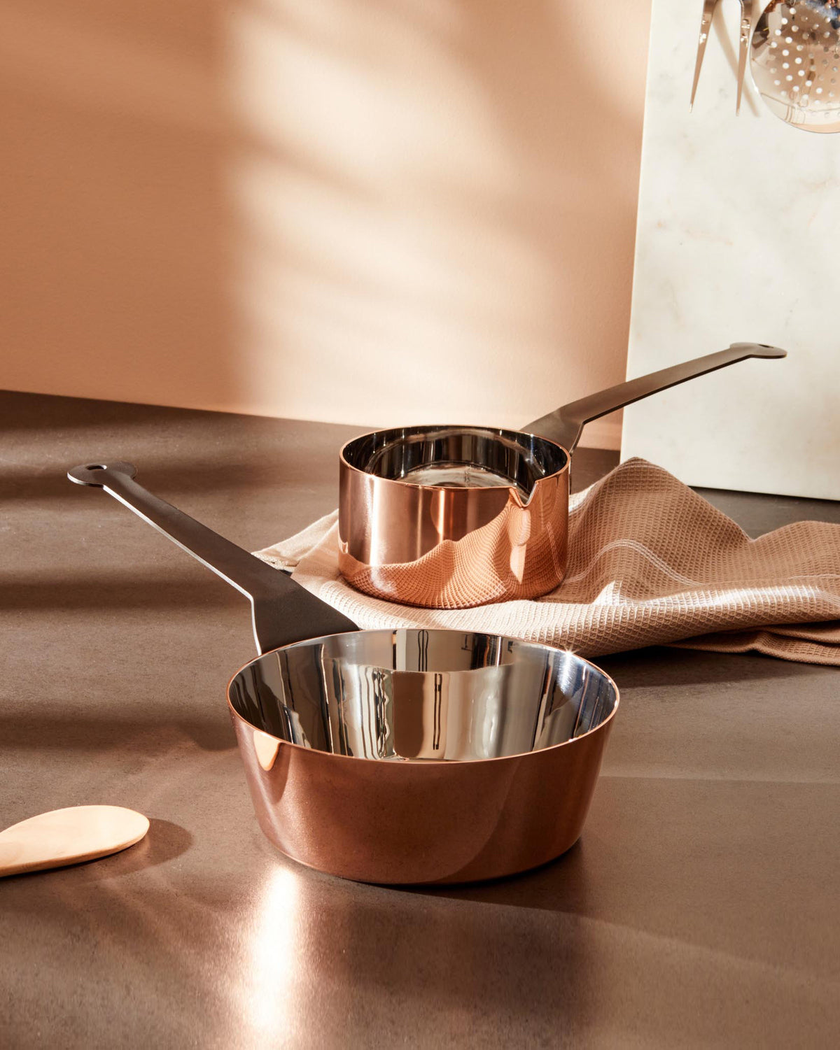 Two shiny copper saucepans by Alessi with matte black handles, one smaller with a pouring spout, on a dark kitchen counter.