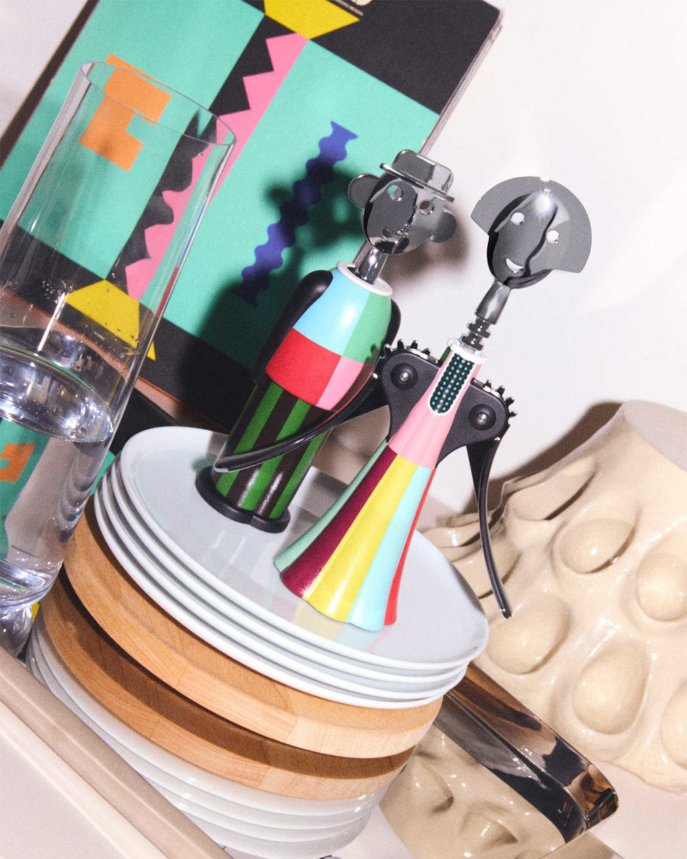 Metallic-headed colorful striped Anna G. corkscrew by Alessi, standing beside a green striped corkscrew on stacked white plates.
