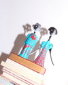 Turquoise Anna G. corkscrew by Alessi with red patterns, standing with a male-figure corkscrew on a stack of books.
