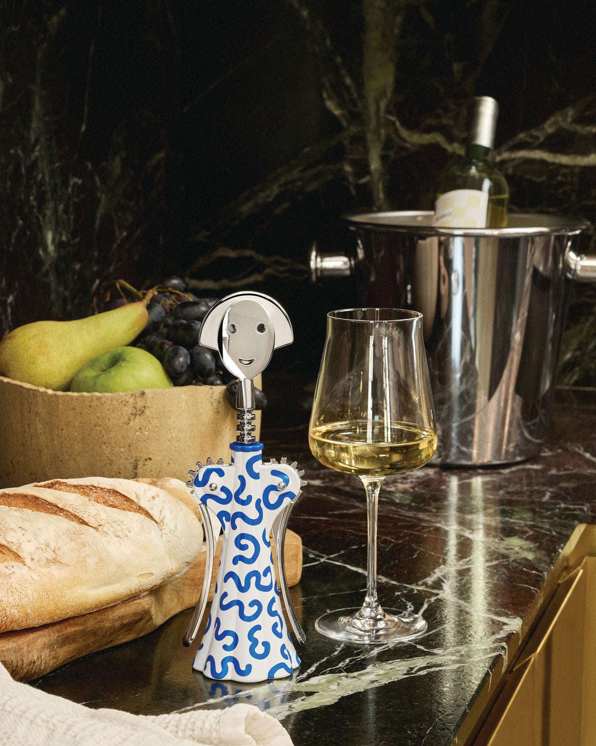 White and blue patterned corkscrew by Alessi with a shiny silver smiling face top, standing upright next to a wine glass.