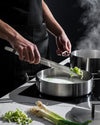 Stainless steel Convivio kitchen tongs by Alessi in use, adding chopped green vegetables to a pot.