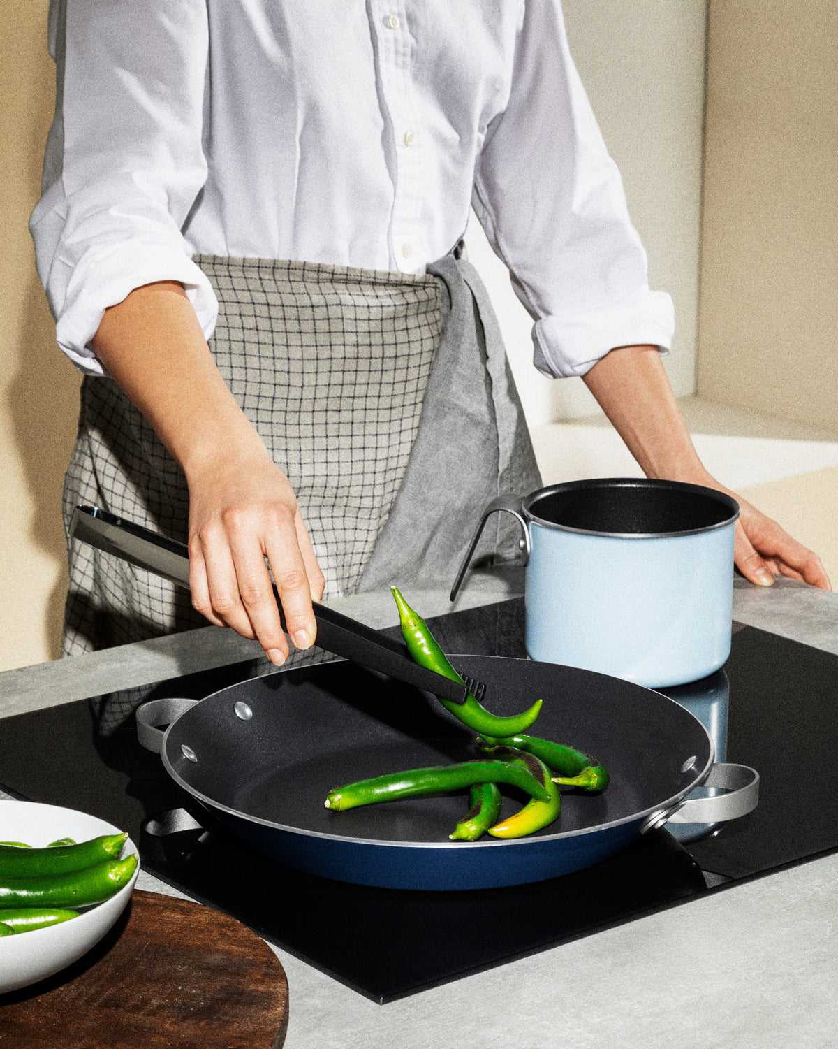 Black and silver kitchen tongs by Alessi gripping green peppers in a blue pan on a stovetop.