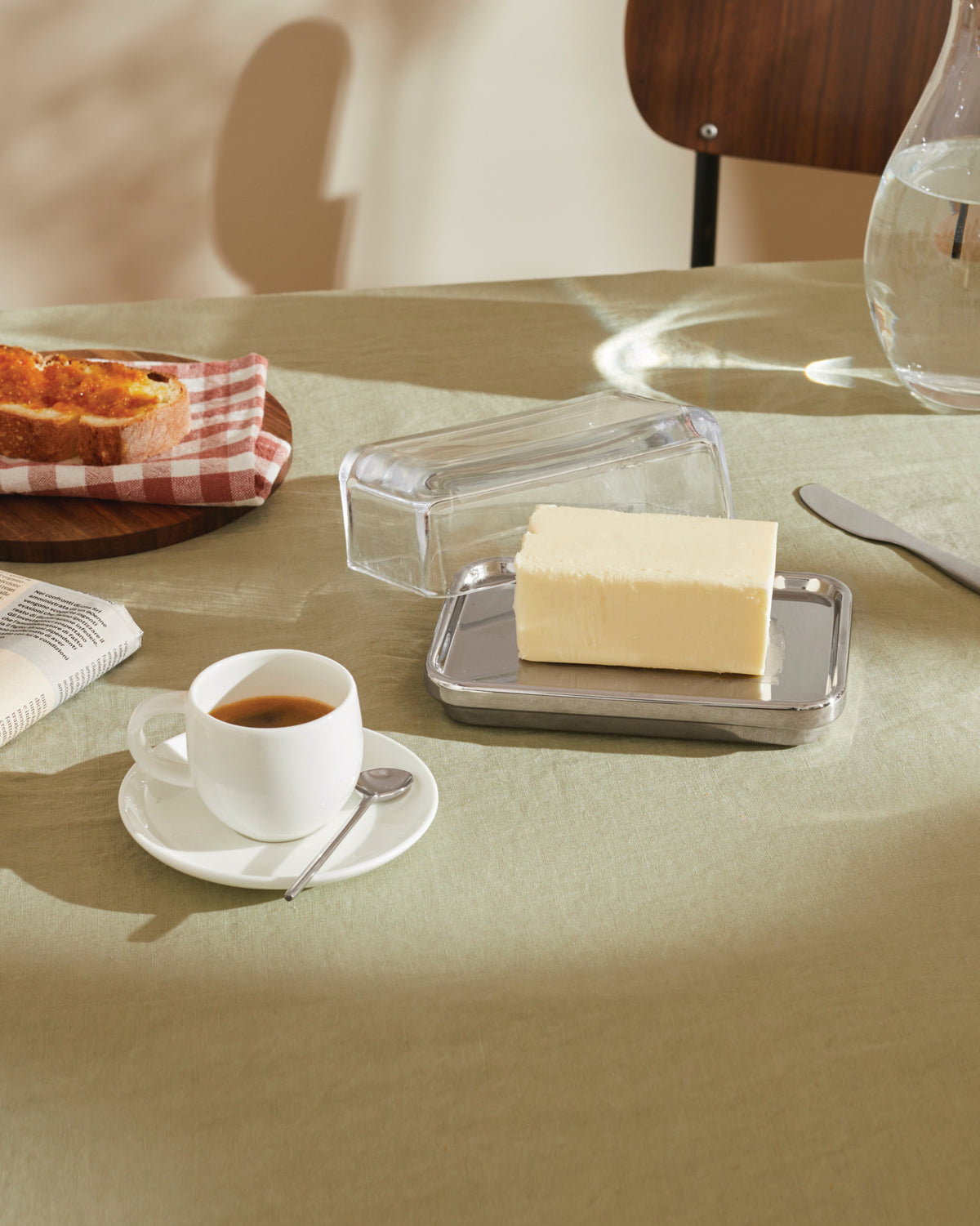 Clear glass and stainless steel butter dish by Alessi holding a block of butter on a light green table.