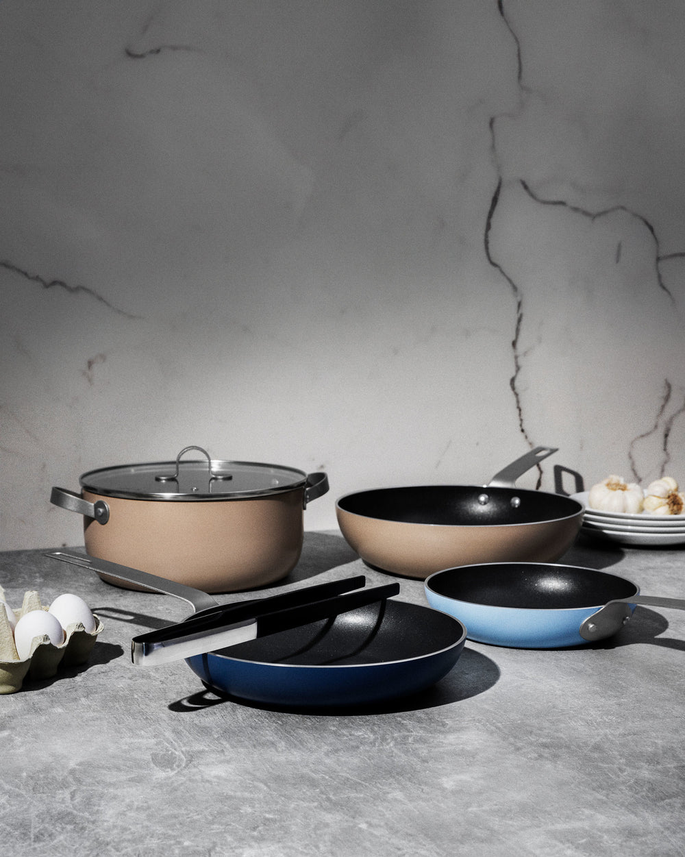 Various non-stick beige and blue pots and pans by Alessi displayed on a grey countertop.