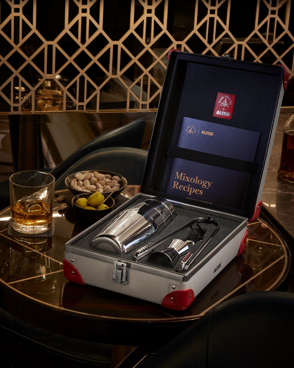 Silver cocktail shaker set by Alessi in an open grey suitcase with red accents, on a glossy dark wood bar table, beside a whiskey glass and snacks.