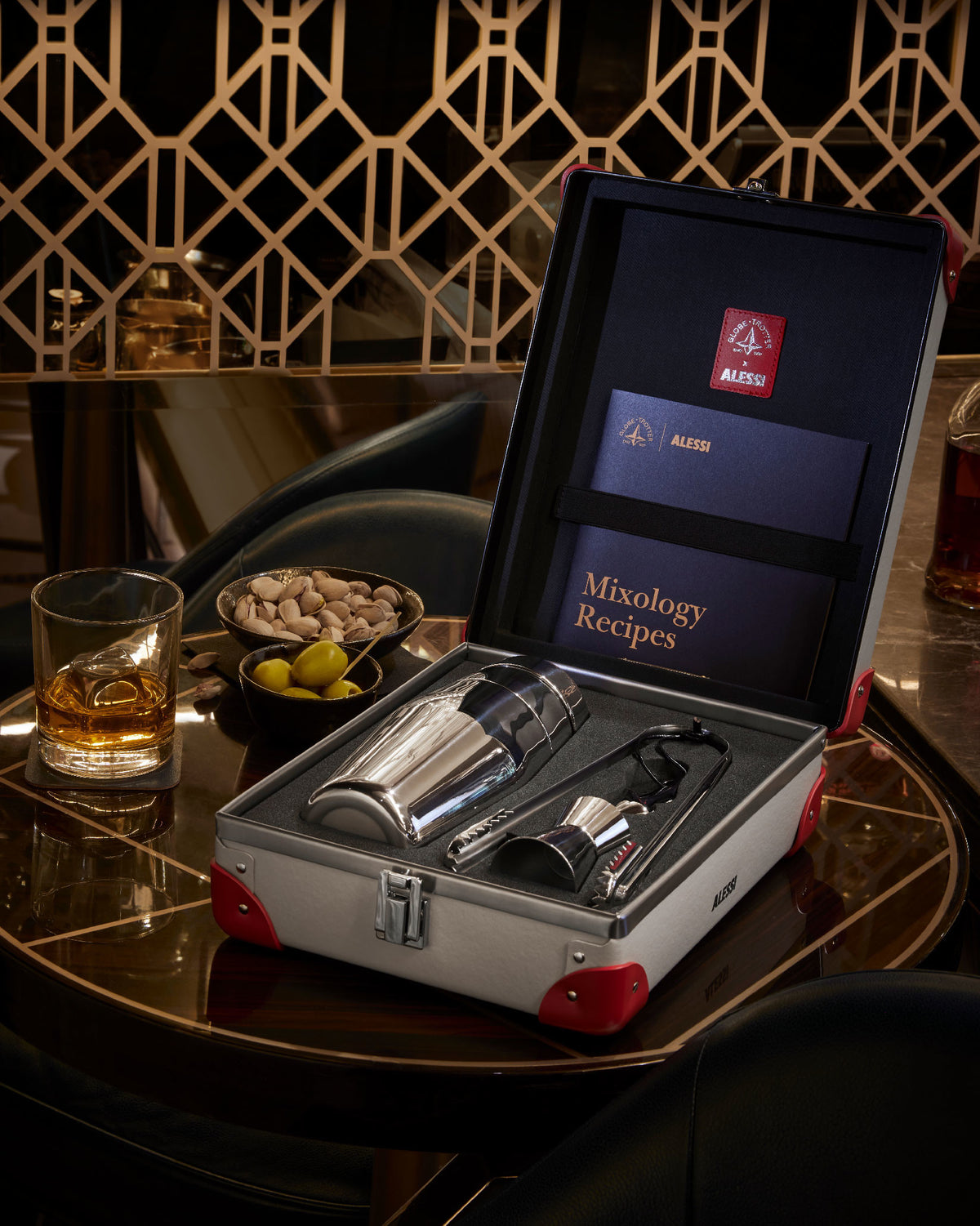 Silver cocktail shaker set by Alessi in an open grey suitcase with red accents, on a glossy dark wood bar table, beside a whiskey glass and snacks.