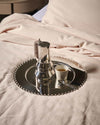 Silver metallic round tray by Alessi with a scalloped edge, holding a Moka pot and espresso cup on pink sheets.