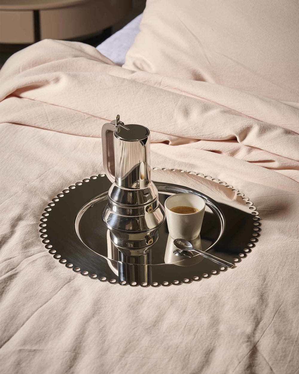 Silver metallic round tray by Alessi with a scalloped edge, holding a Moka pot and espresso cup on pink sheets.