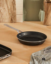 Black non-stick pan by Alessi with a silver handle, on a wooden counter, next to a striped towel and grill pan.