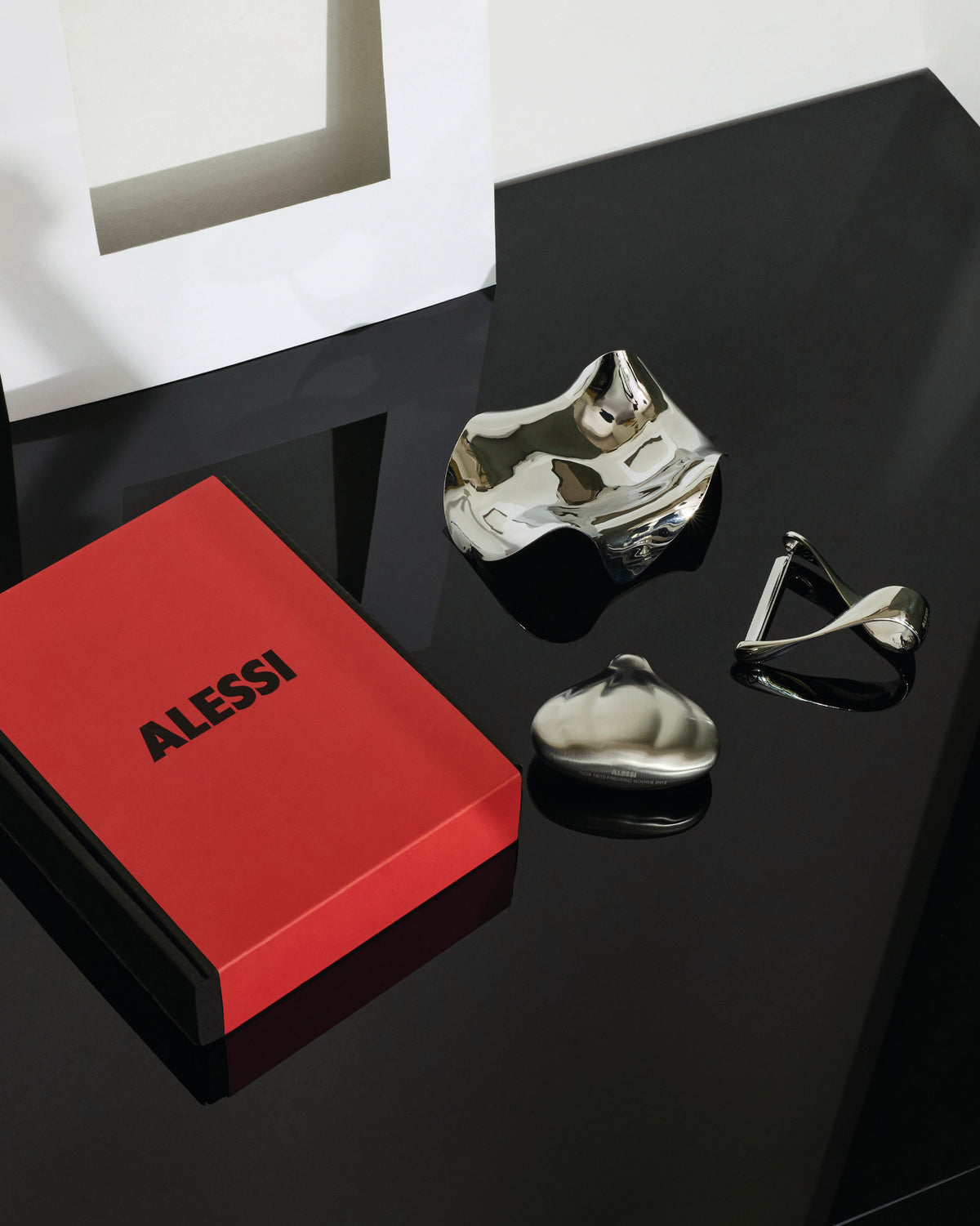 Red gift set with three silver metallic objects by Alessi, including a book-like box and sculptural pieces on a reflective black surface.