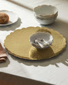 Gold metal tray by Alessi with an ornate scrolled pattern and scalloped edge, set on a white tablecloth.