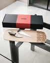 Black and red cheese gift set by Alessi with two silver tools and a carved wooden board.