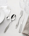 Sleek, polished stainless steel cutlery set by Alessi, with a fork, knife, and two spoons, set on a white table with glasses and plates.