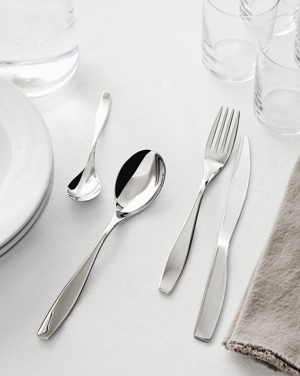 Sleek, polished stainless steel cutlery set by Alessi, with a fork, knife, and two spoons, set on a white table with glasses and plates.