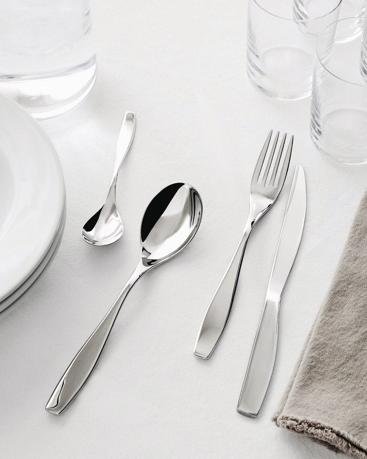Sleek, polished stainless steel cutlery set by Alessi, with a fork, knife, and two spoons, set on a white table with glasses and plates.