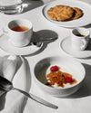 White small bowl by Alessi, filled with yogurt, granola, and fresh raspberries, amidst a breakfast setting.