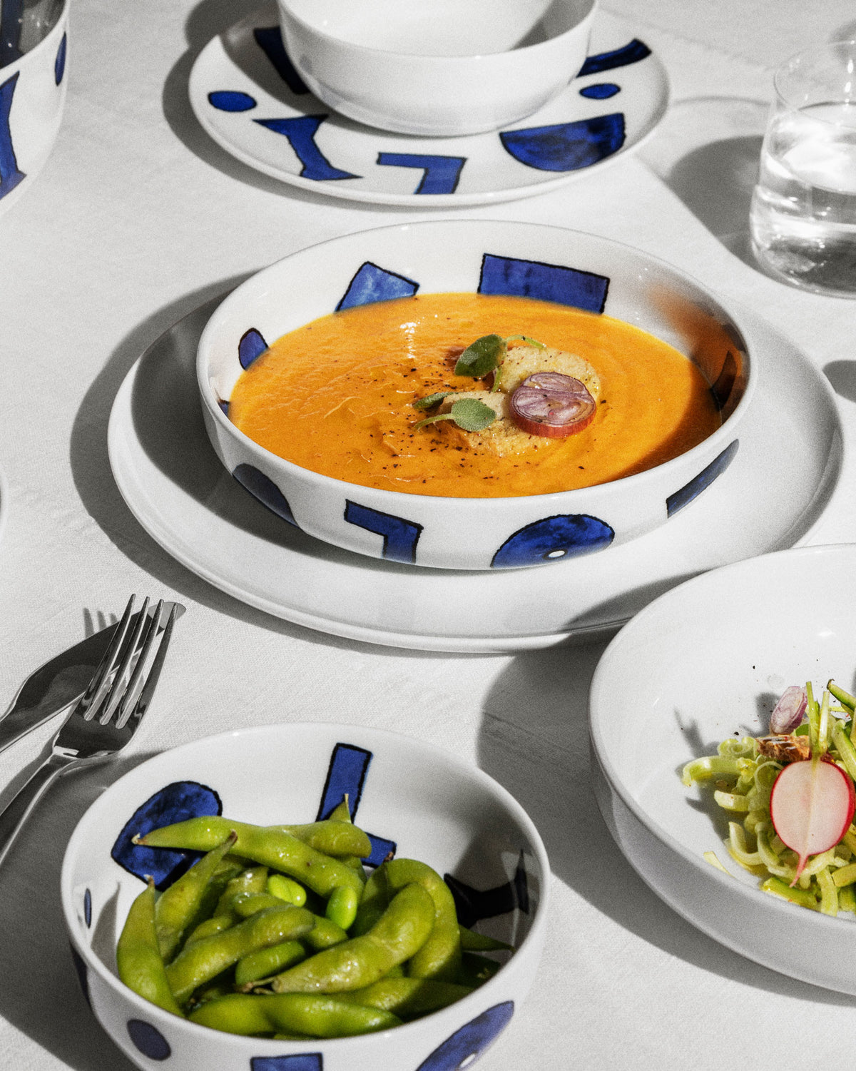 White tableware set with blue geometric patterns by Alessi, displaying soup, edamame, and salad on a white tablecloth.