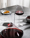 Clear red wine glass by Alessi, with a decanter, an empty glass, and snacks on a grey table.