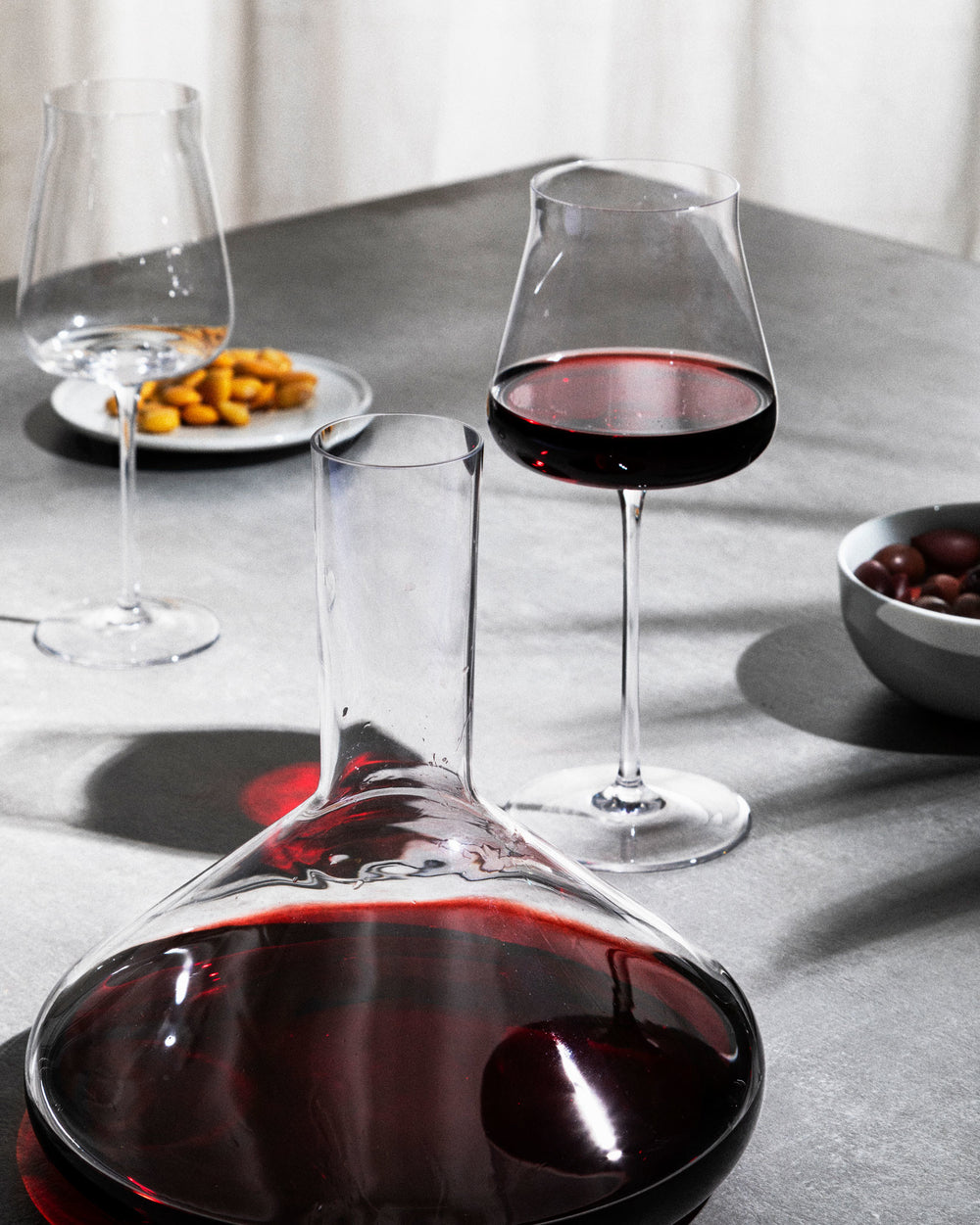 Clear red wine glass by Alessi, with a decanter, an empty glass, and snacks on a grey table.