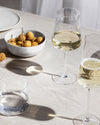 A clear stemmed white wine glass by Alessi, filled with pale wine, displayed with a champagne flute, water glass, and longan fruits on a linen table.