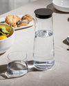 Clear glass pitcher with a black lid by Alessi, filled with water, alongside a drinking glass and bread rolls.