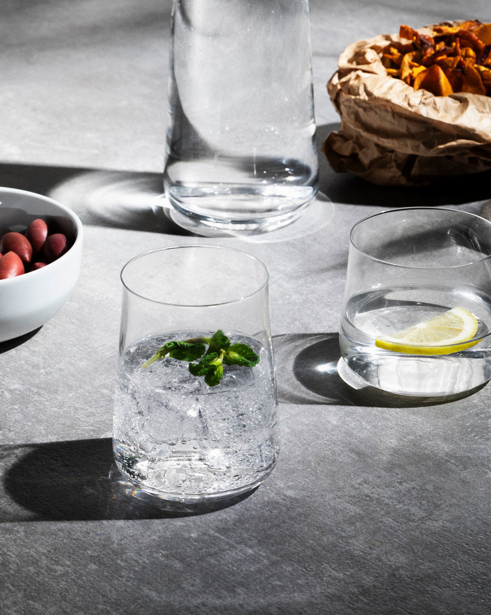 Clear glass Eugenia long drink glasses by Alessi, one with mint, another with lemon, on a gray stone counter.