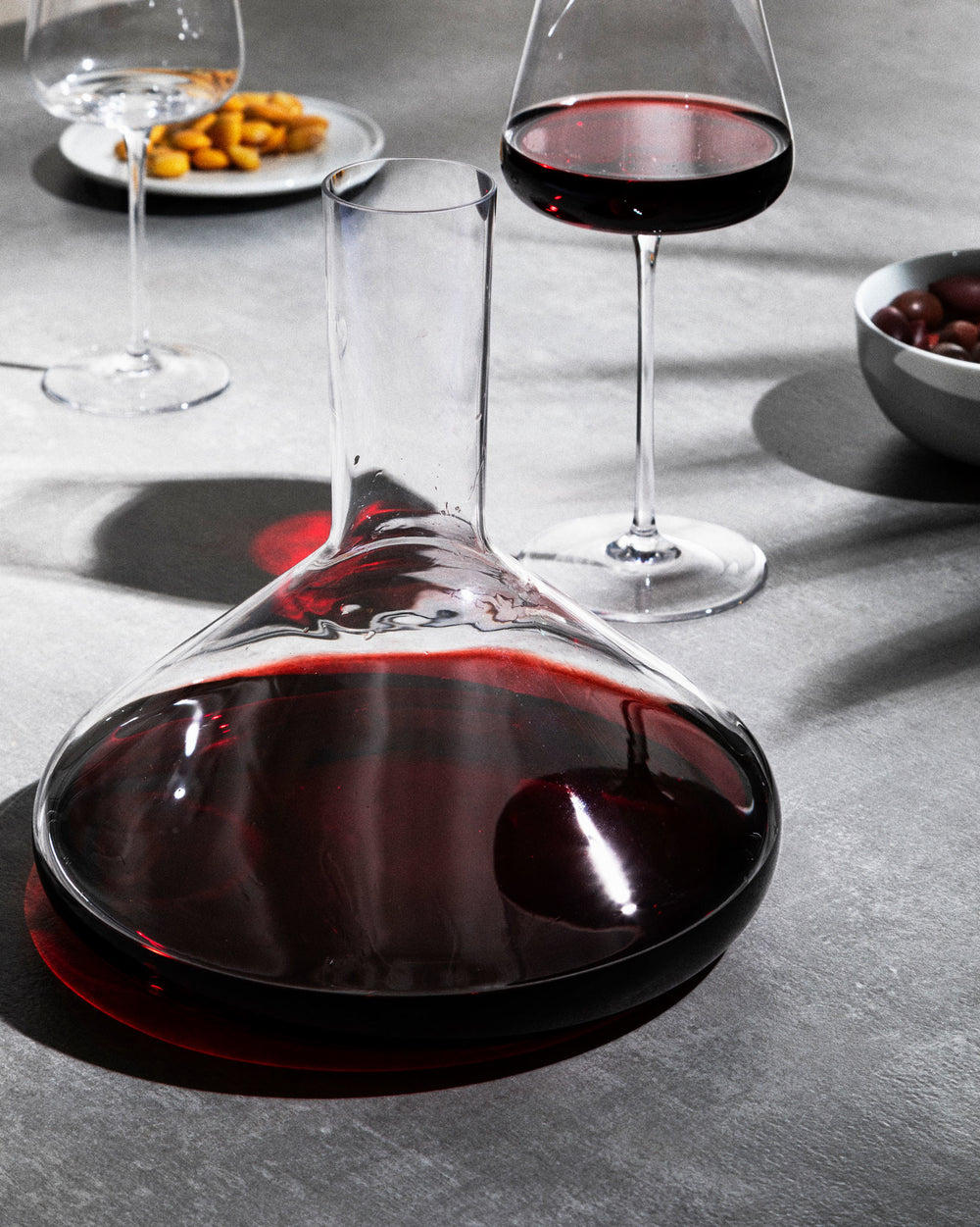 Glass decanter filled with red wine by Alessi, on a grey surface with wine glasses and snacks.