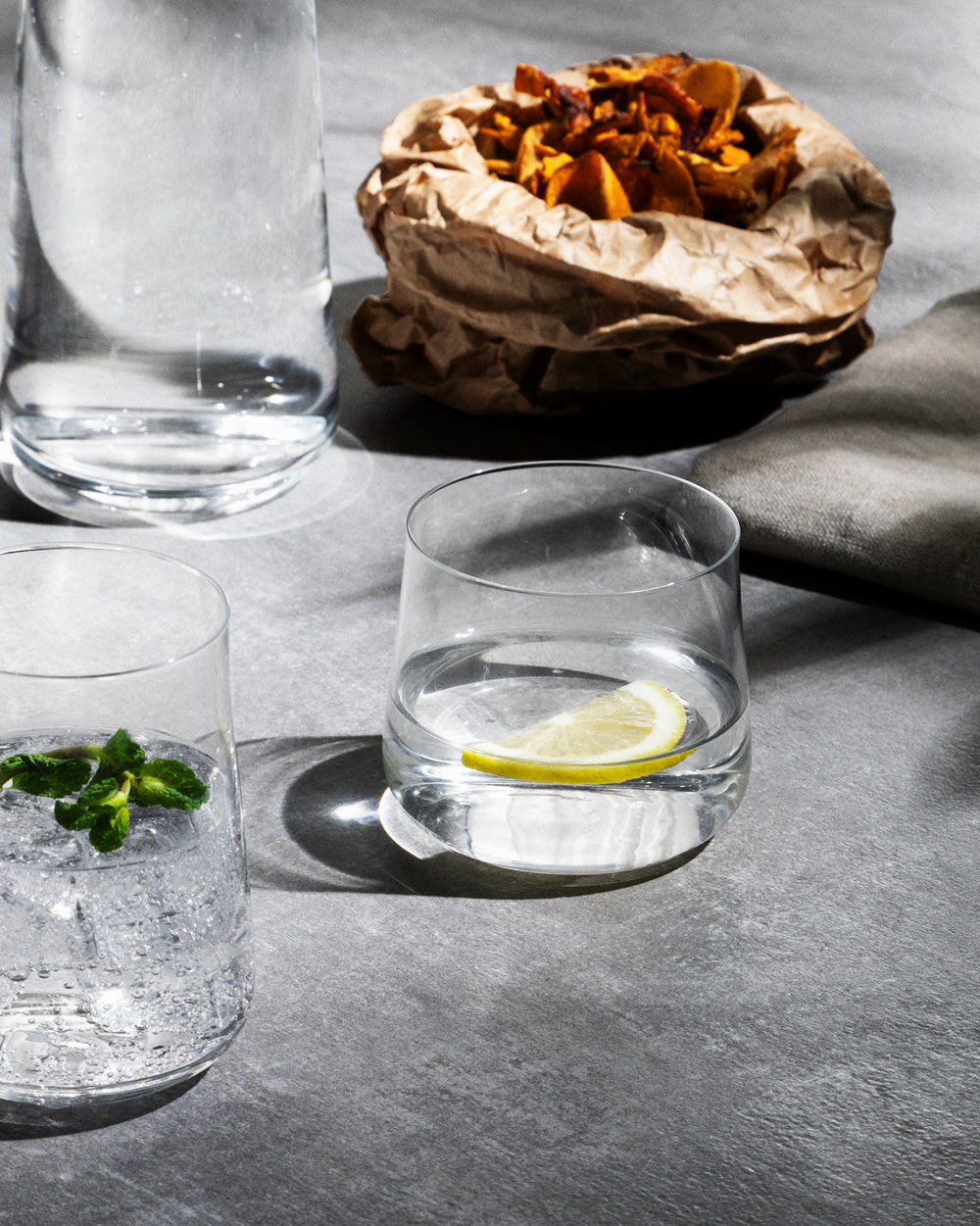 Clear glass water glasses by Alessi, with one holding a lemon slice and another mint, on a grey surface with snacks.