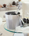 A multi-faceted stainless steel wine cooler by Alessi with water droplets, holding a champagne bottle and ice on a glass table.