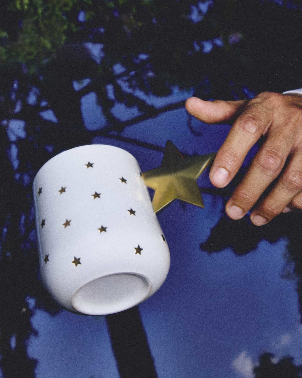 White mug by Alessi with gold star pattern and a shiny star-shaped handle on a blue reflective surface, a hand reaching for it.