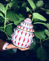 White and red Christmas tree container by Alessi with gold trim and star topper, held in hand amidst green leaves.