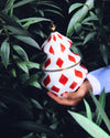 White and red ceramic Christmas tree decoration by Alessi, featuring a diamond pattern and a gold top, held in a hand amidst green leaves.