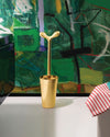 Gold toilet brush by Alessi with a distinctive 'Y'-shaped handle, standing in its holder against an abstract green painting.