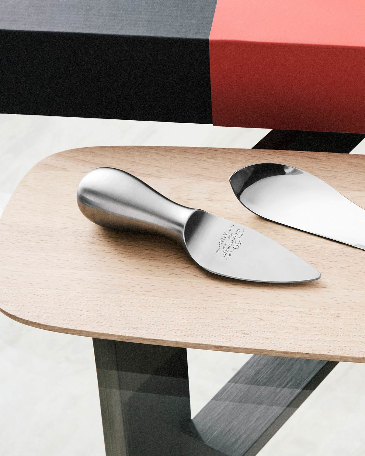 Sleek, brushed stainless steel cheese server by Alessi, engraved with '50 IL CONSORZIO 1954~2004 ANNI', on a light wood board.