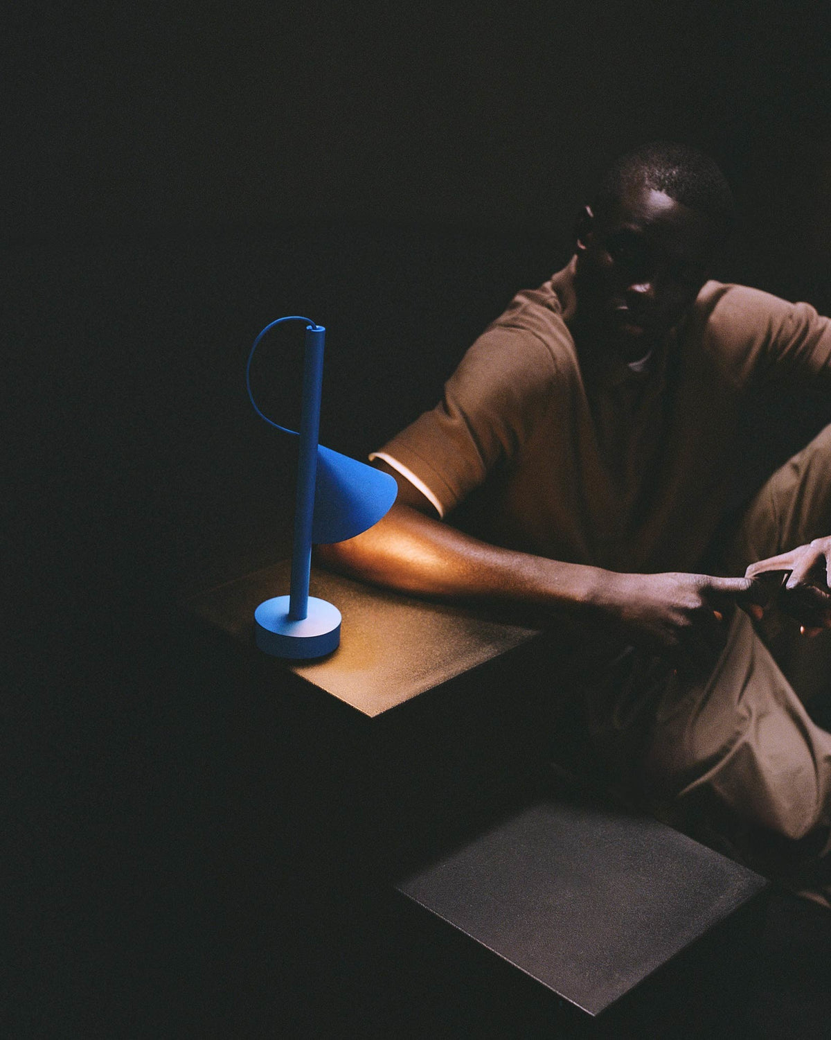 Blue table lamp by Alessi with a minimalist design, casting a warm glow on a man and a gold surface.