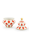 White ceramic decorative container by Alessi with red diamond patterns, gold trim, and a separate lid featuring a gold finial.
