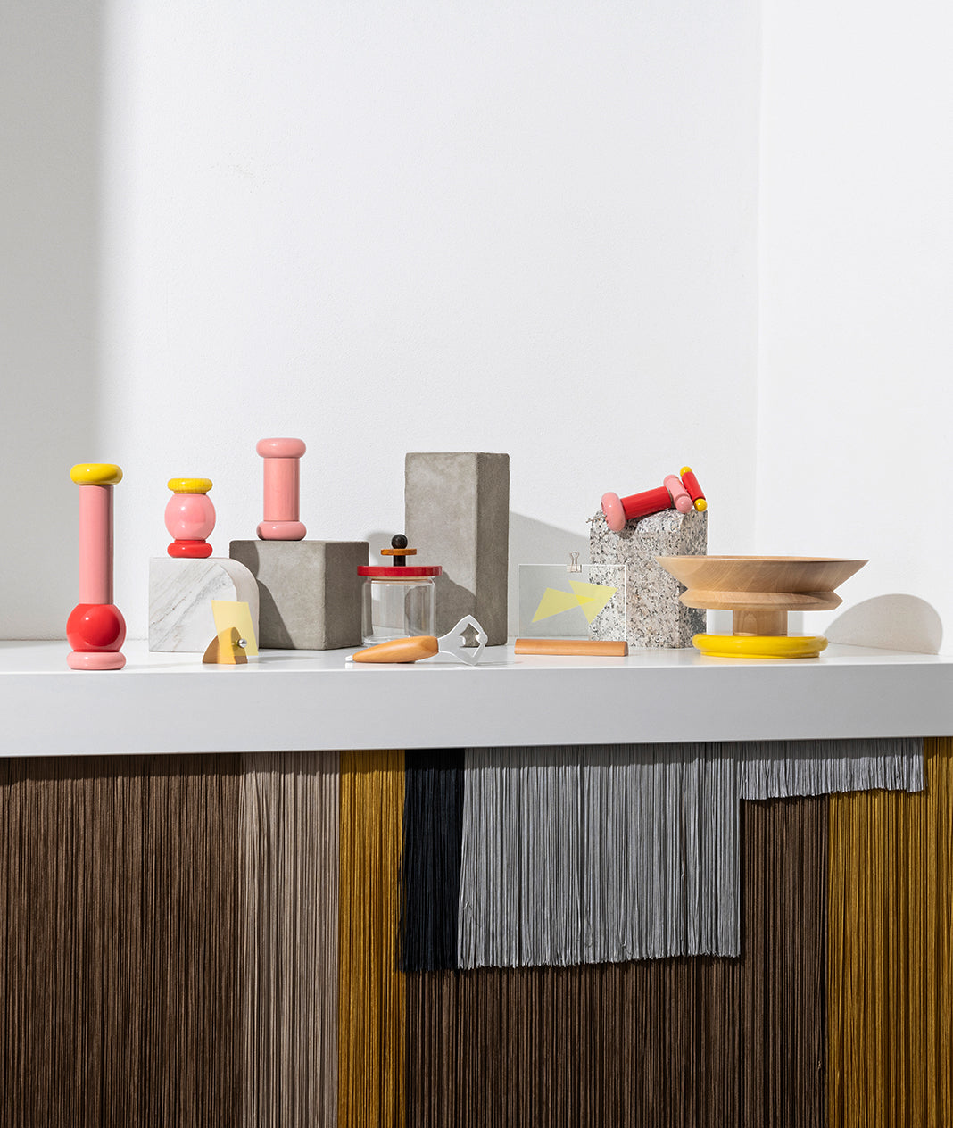 Colorful wooden and concrete objects arranged on white shelf with fringed fabric below.