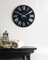 A round black wall clock with white Roman numerals by Alessi, displayed on a white textured wall above a black fireplace mantel.
