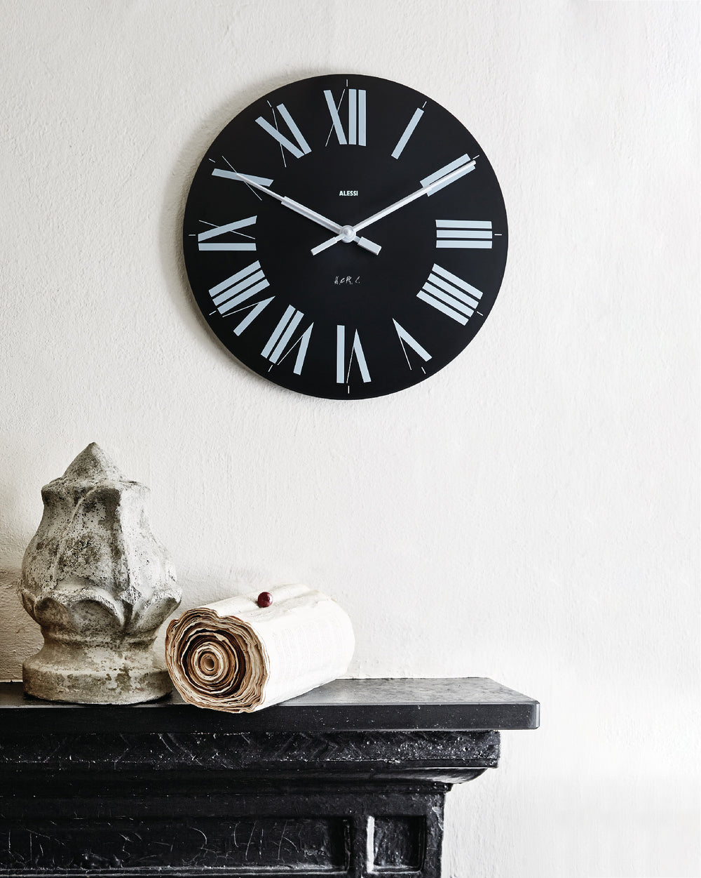 A round black wall clock with white Roman numerals by Alessi, displayed on a white textured wall above a black fireplace mantel.