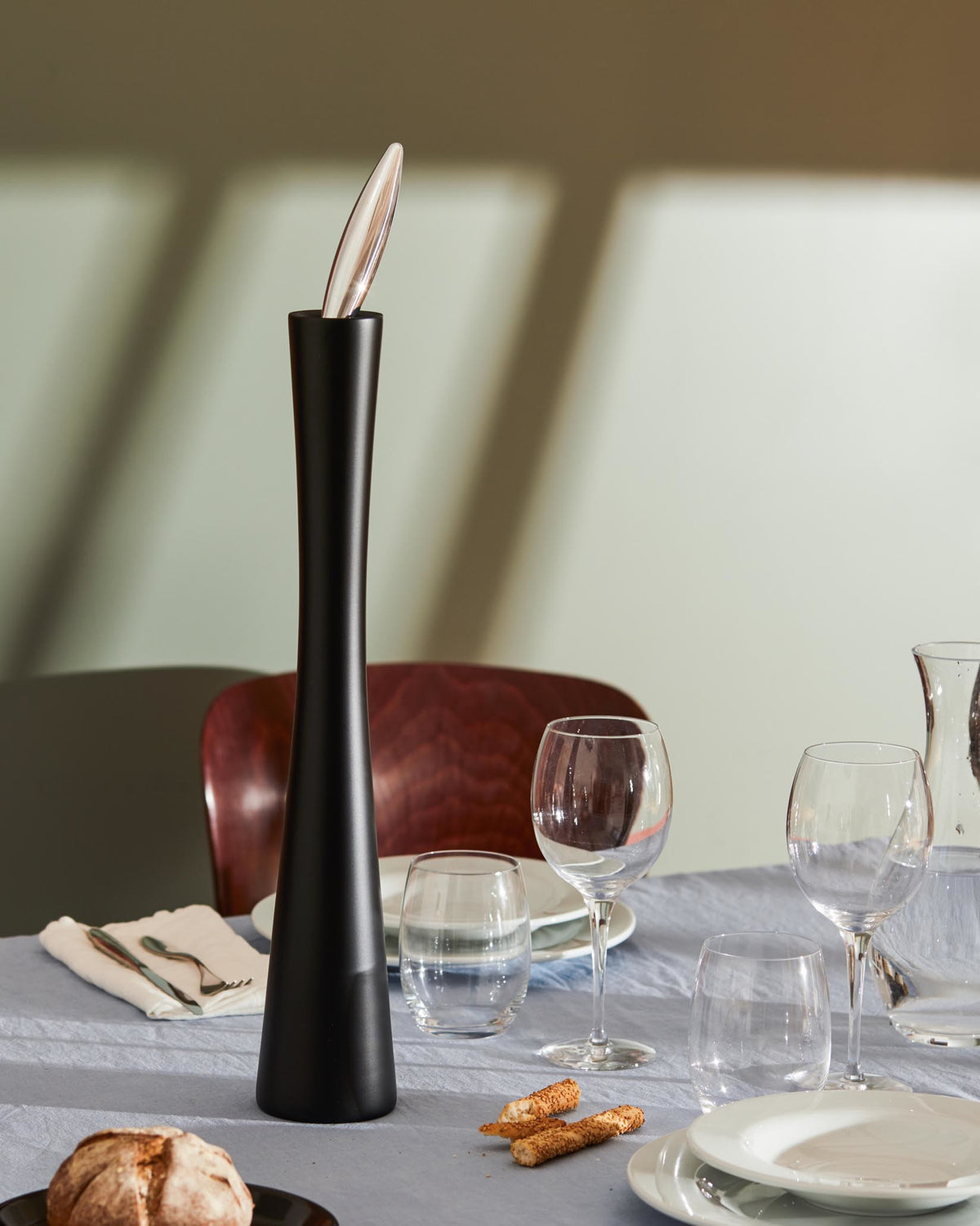 Tall matte black grinder by Alessi with a clear leaf-shaped top, styled on a dining table.