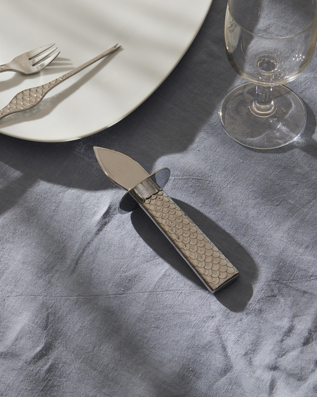 Silver oyster knife by Alessi with a fish scale patterned handle, resting on a blue linen tablecloth.