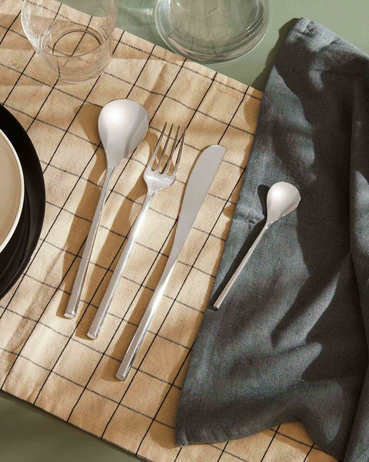 Silver metal flatware set by Alessi, including a soup spoon, fork, knife, and teaspoon, displayed on a plaid placemat and linen napkin.