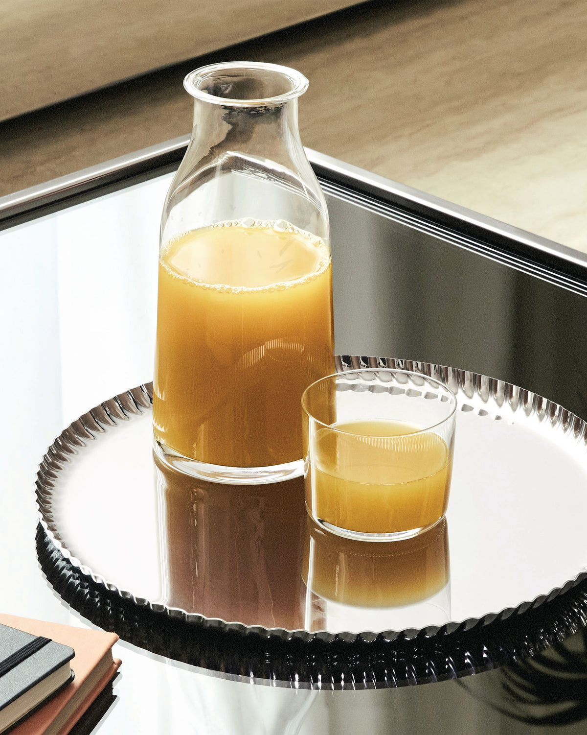 Silver oval tray by Alessi with a ruffled edge, holding a bottle and glass of orange juice on a mirrored table.